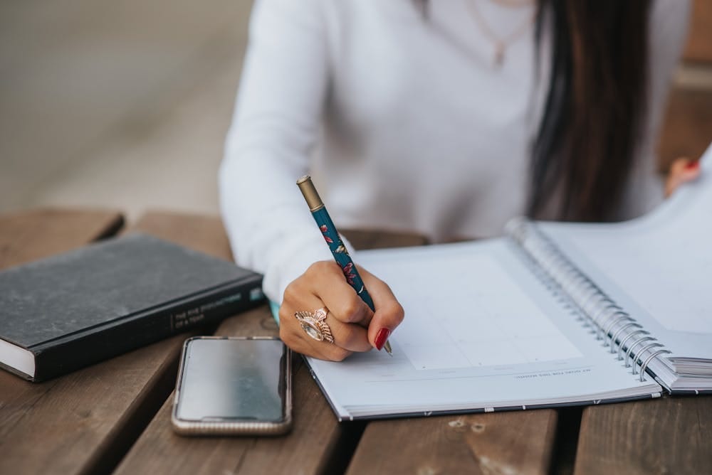 A woman sitting at a wood table taking notes in a notebook.